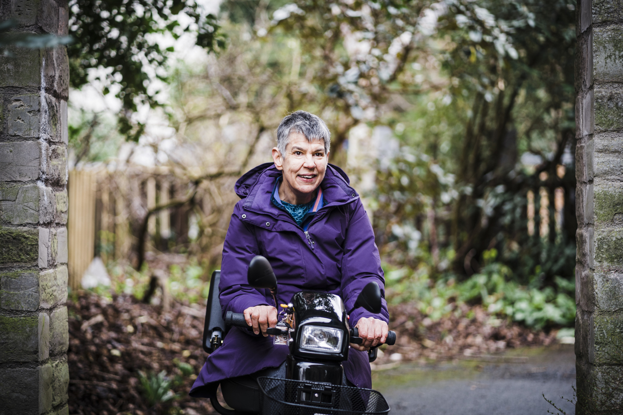 Wearing a purple winter coat, Annalu drives her motorized wheelchair through a stone gate on the wooded campus of Dundee University.
