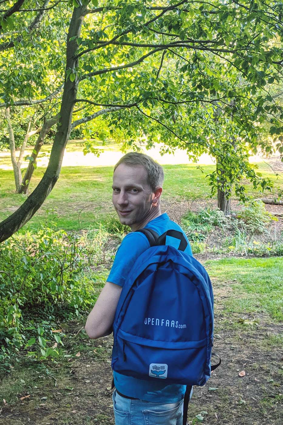 Photo of Alex Ellis surrounded by greenery, wearing an OpenFaaS branded backpack.