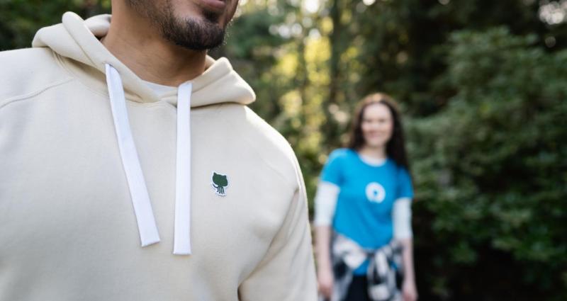 A photo of someone wearing the GitHub Sportiqe hoodie with a green invertocat patch. In the background is someone wearing the fitted invertocat blue tee.